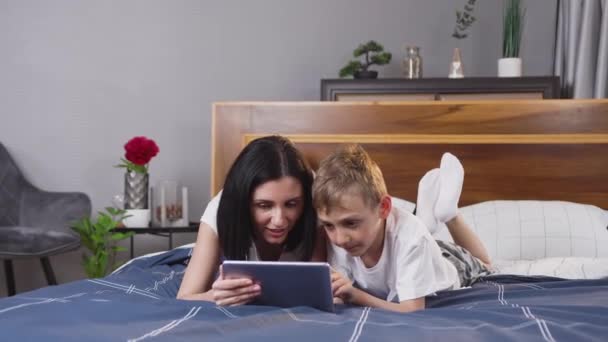 Charmante brune de 30 ans à l'esprit aigu couchée sur un lit confortable avec son beau fils gai et utilisant une tablette PC — Video