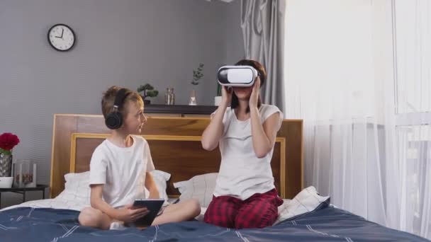 Concept familial où beau petit garçon joyeux utilisant tablette PC tandis que sa belle mère souriante insouciante en utilisant casque de réalité virtuelle à la maison — Video