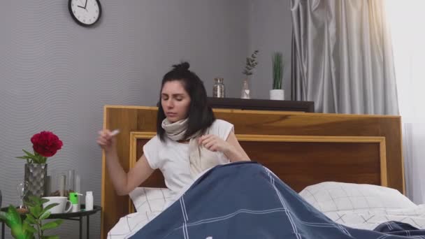 Upset sick dark-haired woman with scarf around her neck measuring temperature using thermometer and drinking tea during flu disease at home — Stock Video