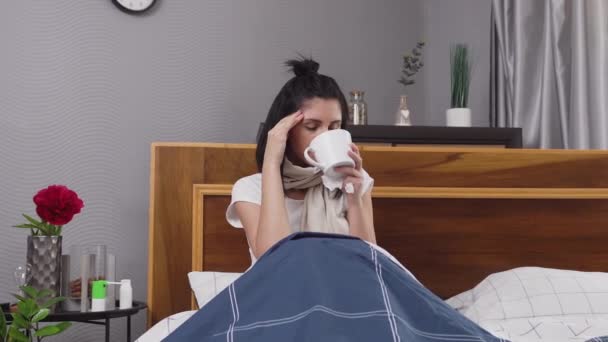 Vista frontal de la mujer joven enferma molesta que se sienta en la cama del dormitorio y beber té mientras se trata en casa como resfriado con tos y dolor de cabeza — Vídeos de Stock