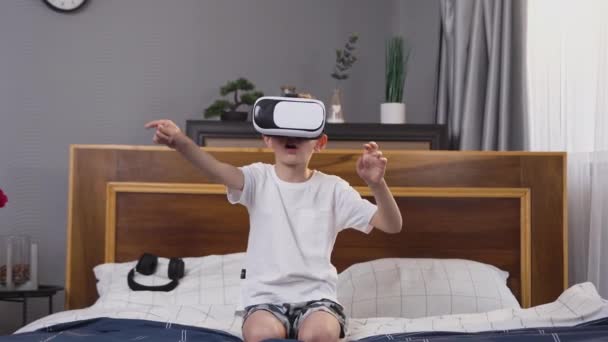 Schöner kleiner Junge im Virtual-Reality-Headset sitzt auf dem Bett und spielt Spiel auf imaginärem Bildschirm — Stockvideo