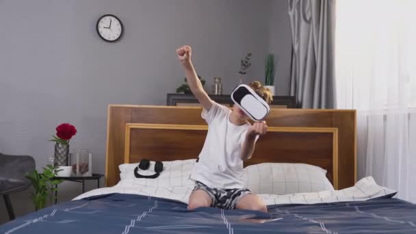 Atraente emocional positivo menino pequeno em óculos 3d especiais jogando corrida de vídeo, sentado na cama no quarto — Vídeo de Stock