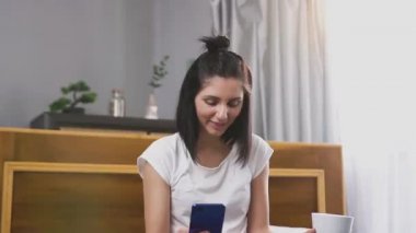 Siyah saçlı, gülümseyen genç bir kadın telefonda komik videolar izliyor ve büyük pencerenin yanında çay içiyor.