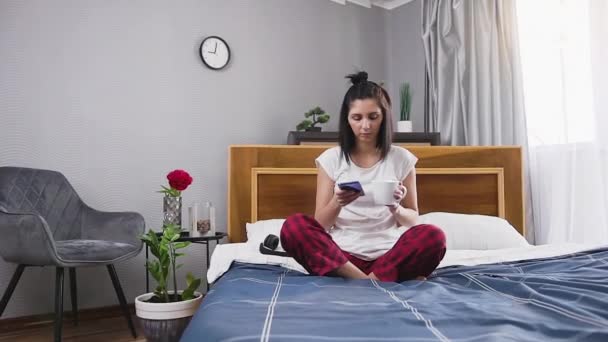 Вид спереду задоволений щасливою молодою брюнеткою, яка сидить на спальні та п'є чай під час використання телефону — стокове відео