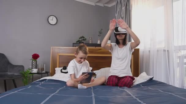 Guapo sonriente feliz niño de 8 años con i-pad sentado en la cama cerca de su encantadora madre alegre en auriculares de realidad virtual — Vídeo de stock
