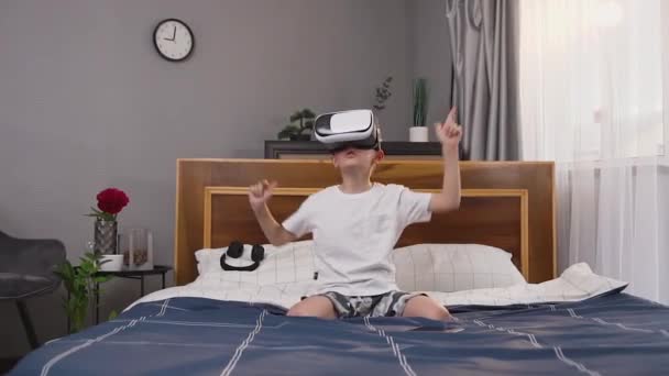 Portret van knappe emotionele positieve kleine jongen in huiskleding die zitten op bed en videospel spelen met behulp van augmented reality bril — Stockvideo