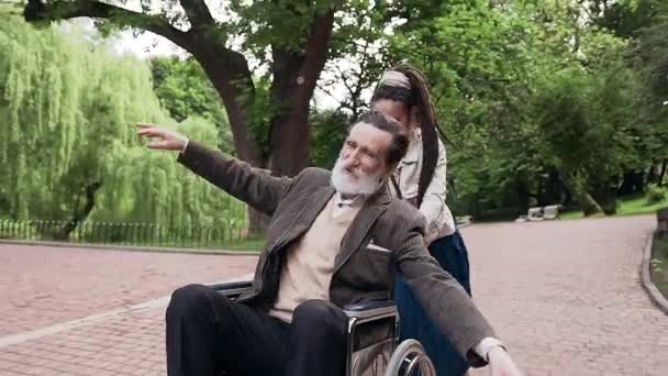 Özgürlük duygusu, tekerlekli sandalyedeki gülümseyen sakallı emeklinin neşeli ve mutlu torunuyla şehir parkında rastalı saçlarıyla eğlenmesi. — Stok video