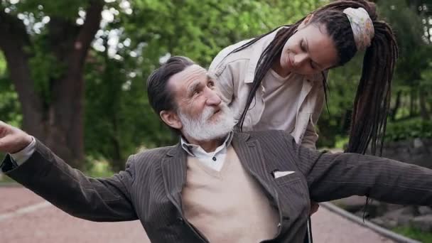 Likable satisfied high-spirited young woman with dreadlocks enjoying leisure with her respected positive smiling bearded granddad in wheelchair in green park — Stock Video