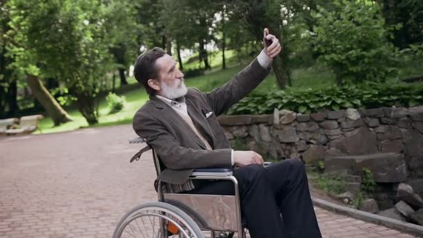 Probable positif satisfait barbu homme âgé en fauteuil roulant photographie sur smartphone dans un parc vert, au ralenti — Video