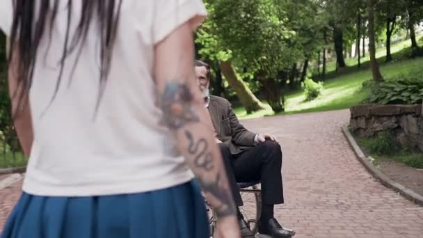Hezký úsměv přátelský vousatý starý muž na invalidním vozíku objímání jeho moderní krásné vnučky s dredy v parku během chůze — Stock video