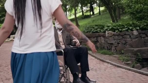 Сімейна концепція, де задоволений усміхнений люблячий зрілий чоловік у інвалідному візку з сірою бородою зустріч з обіймами його красивою сучасною молодою онукою з дредлоками в парку — стокове відео