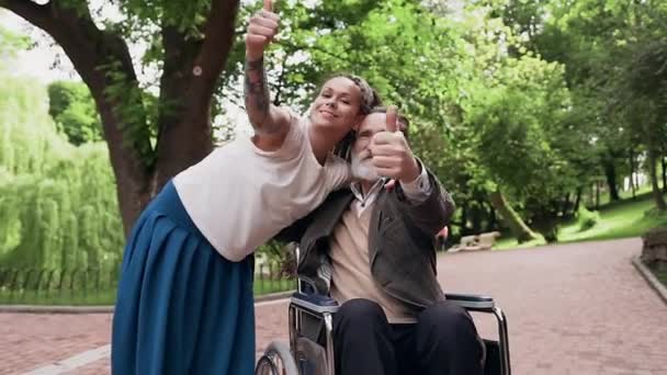 Bonne humeur amicale positive petite-fille chanceuse et grand-père barbu en fauteuil roulant regardant la caméra avec des sourires et faire des gestes OK dans le parc — Video