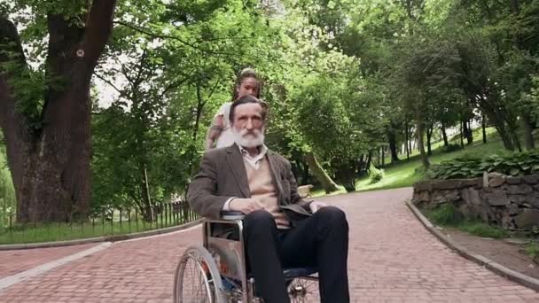 Retrato de boa aparência feliz amorosa moderna menina de 25 anos que se divertindo junto com seu respeitado avô barbudo satisfeito em cadeira de rodas no parque — Vídeo de Stock