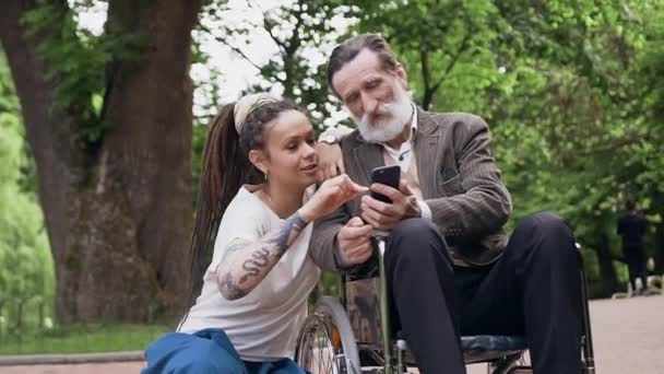 Portrait attrayant de sympathique joyeuse petite-fille avec dreadlocks et respecté grand-père à la retraite barbu en fauteuil roulant en utilisant smartphone dans le parc urbain — Video