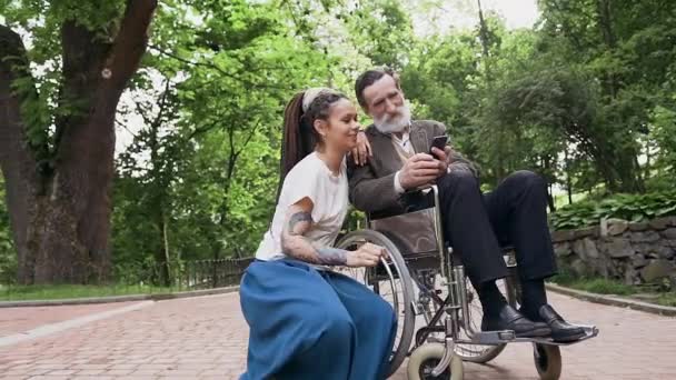 Slow motion van tevreden goedgehumeurd trendy meisje met dreadlocks op zoek naar smartphone samen met haar goed uitziende bebaarde 70-jarige grootvader in prachtig park — Stockvideo