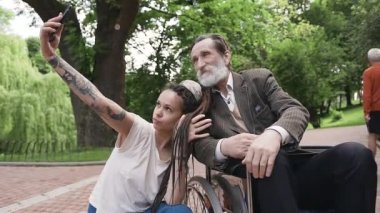 Parkta tekerlekli sandalyede oturan yakışıklı, pozitif sakallı büyükbabasıyla selfie çeken enfes, modaya uygun, modern genç kızın portresi.