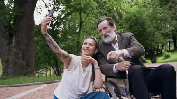 Charming riang tersenyum cucu modern dengan gimbal membuat selfie bersama-sama dengan kakek berjenggot dihormati di kursi roda di taman hijau selama berjalan — Stok Video