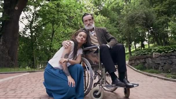 Good-tempered lucky carefree trendy woman with dreadlocks and her sedentary contented respected bearded old granddad in wheelchair looking at camera in urban park — Stock Video