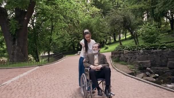 Atraente positivo despreocupado moderna neta com dreadlocks sair empurrando cadeira de rodas com seu sorrindo estimado avô idoso barbudo no parque verde — Vídeo de Stock