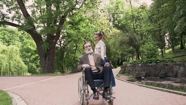 Goed uitziende vrolijke moderne jonge vrouw met dreadlocks brengen gezamenlijke vrije tijd met haar gewaardeerde zorgeloze volwassen baard opa in groen park — Stockvideo