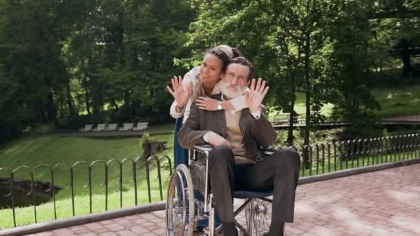 Bonito positivo respeitado barbudo homem de 70 anos em cadeira de rodas e encantadora menina moderna sorridente com dreadlocks posando na câmera com as mãos acenando no parque verde — Vídeo de Stock