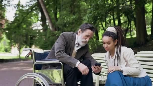 Familienkonzept, wo geschätzte sitzende alte bärtige Mann im Rollstuhl zeigt etwas seine attraktive moderne selbstbewusste junge Enkelin mit Dreadlocks auf Handy im grünen Park — Stockvideo