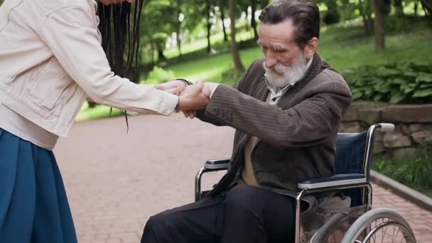 Concepto de familia donde encantador feliz sonrisa chica moderna con peinado de moda ayudando a levantarse de la silla de ruedas su hermoso abuelo alegre y despreocupado en el parque verde — Vídeo de stock