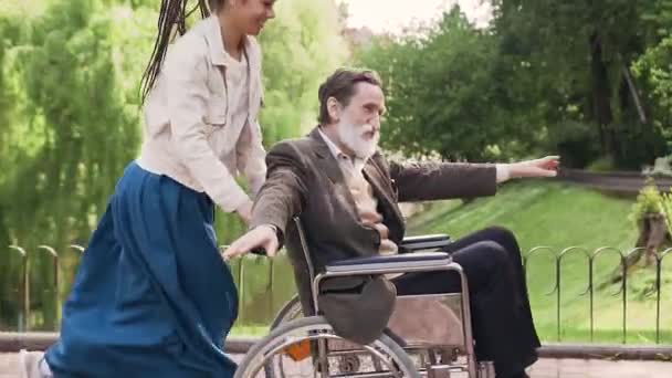 Fröhliches, gut aussehendes, unbeschwertes modernes Mädchen mit Dreadlocks und älterer behinderter Mann im Rollstuhl, der beim Spazierengehen im grünen Park gemeinsam Spaß hat — Stockvideo