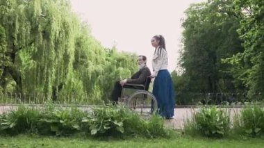 Modern saç stiline sahip, çekici ve modaya uygun bir genç torunun portresi. Park sokağında oturan saygıdeğer büyükbabasıyla tekerlekli sandalye itiyor.