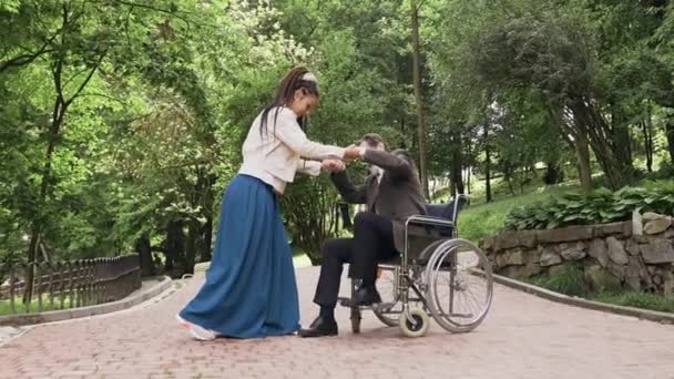 Adorable amante joven feliz con rastas ayudando a levantarse de la silla de ruedas su estimado apuesto abuelo barbudo maduro en el parque verde, 4k — Vídeo de stock