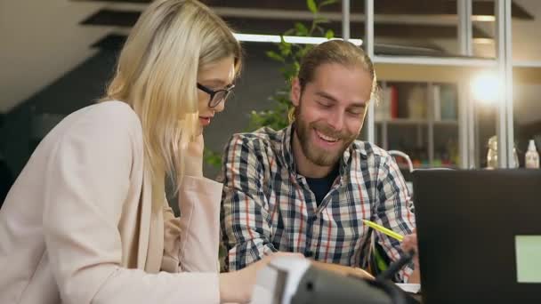 Mouvement lent d'un bel homme barbu souriant qui parle avec une blondinette assez satisfaite dans des lunettes pendant le travail conjoint à la table du bureau — Video