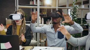 Modern ofis odasında hayali ekran üzerinde çalışan, sanal gerçeklik kulaklığı takmış ciddi ve çok ırklı insanların ağır çekimleri.