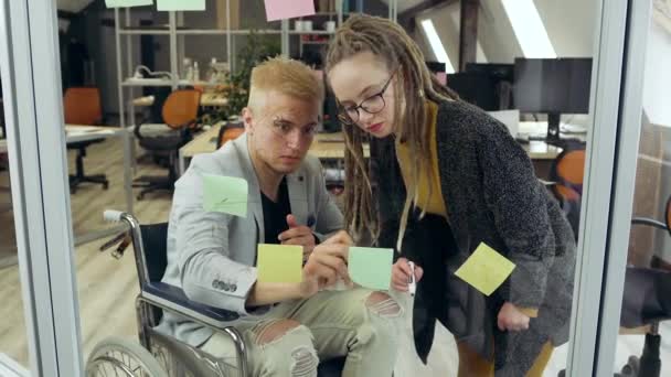 Detailní záběr pohledný světlovlasý podnikavý muž na invalidním vozíku, který diskutuje se svou moderní stylovou kolegyní s dredy nové obchodní strategie pomocí skleněné stěny a lepkavé poznámky v — Stock video