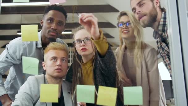 Widok z przodu sympatyczny doświadczony młody wielorasowy zespół start-up, który omawia ich nową strategię, model, który jest przedstawiony na przezroczystej szklanej tablicy w biurze — Wideo stockowe