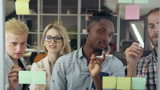 Vooraanzicht van sympathieke goed gehumeurde slimme moderne gemengde ras groep die in de buurt van glazen plank in kantoorruimte staan en bespreken details van business project — Stockvideo