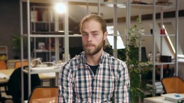 Yakışıklı, kendine güvenen, sakallı genç bir adam. Çağdaş çalışma alanında duruyor ve kameraya bakıyor.