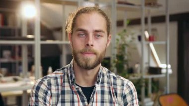 Yakışıklı, kendine güvenen genç sakallı adam çağdaş ofis odasında duruyor ve ciddi bir manzarayla kameraya bakıyor.