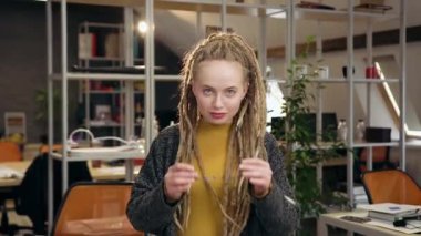 Oldukça olumlu, modern stil sahibi, rastalı gözlüklerini takan ve modern ofis odasında büyüleyici bir gülümsemeyle kameraya bakan genç bir kadın.