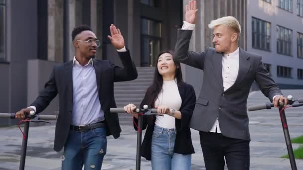 Retrato de alegre amigável jovem de cabelos claros cara, afro-americano e asiático menina que se divertindo juntos durante a caminhada na rua com scooters elétricos e dando alta cinco uns aos outros — Vídeo de Stock
