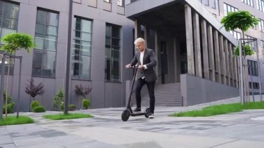 Çekici, hoş, şık, açık renk saçlı genç adam güzel mimari binasının yanında kendi e-scooterıyla geziyor.