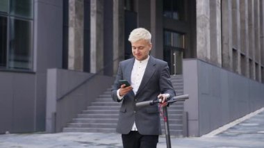 Ekolojik ulaşım konsepti, şık giyinen yakışıklı, mutlu, sarışın bir adamın kendi e-scooterıyla dışarıda yürürken aynı anda telefon kullanması.