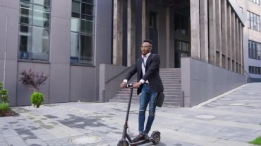 Yakışıklı, kendine güvenen, şık Afrikalı Amerikalı modern binaların yakınındaki şehir sokağında e-scooterla geziyor.