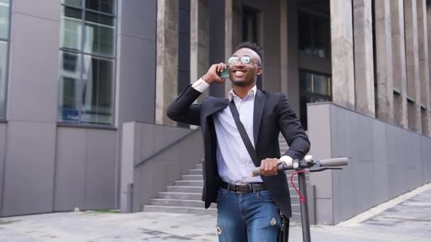 Nahaufnahme attraktiver zufriedener erfolgreicher junger schwarzhäutiger Büroangestellter, der mit seinem eigenen Elektroroller unterwegs ist und im Freien telefoniert — Stockvideo