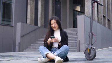 Kabul edilebilir genç Asyalı stil sahibi kız beton zeminde oturuyor güzel şehir binasının arka planında kendi e-scooterının yanında ve akıllı telefonunu kullanıyor.