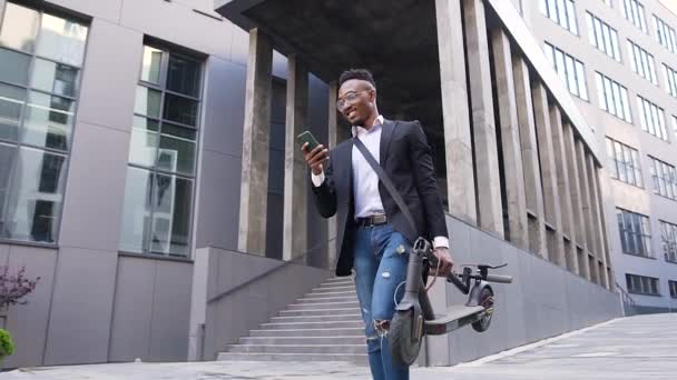 Likable smiling happy young african american in trendy wear walking near beautiful modern building with personal e-scooter in hand and talking on phone — Stock Video