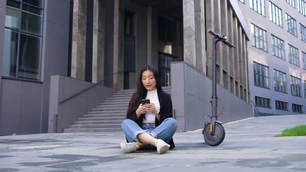 Mooie tevreden glimlachende jonge aziatische meisje zitten op asfalt in de buurt van haar e-scooter op moderne gebouw achtergrond en met behulp van haar telefoon — Stockvideo