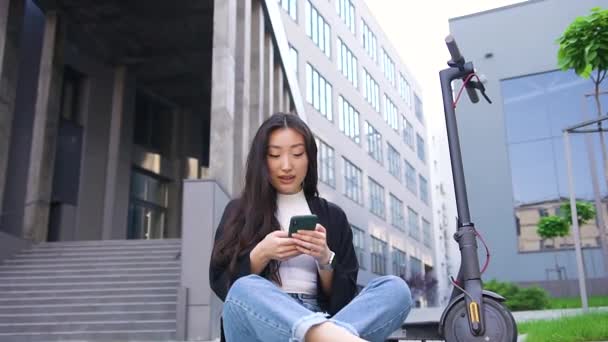 Imádnivaló szerencsés vidám fiatal ázsiai lány alkalmi ruhák ül az aszfalton a város épület hátterében, és használja a telefont — Stock videók