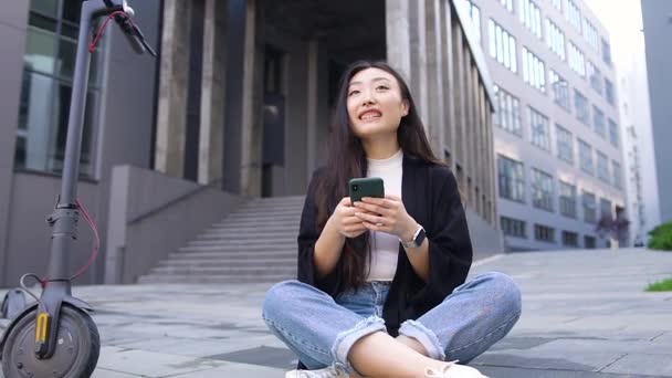 Aantrekkelijk glimlachend moderne jonge Aziatische vrouw zittend op betonnen grond op de achtergrond van het stadsgebouw en met behulp van haar smartphone — Stockvideo