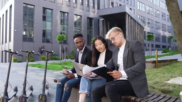 Gros plan de jeunes gens de race mixte positifs attrayants qui sont assis sur le banc près du bâtiment urbain et qui travaillent avec des documents — Video