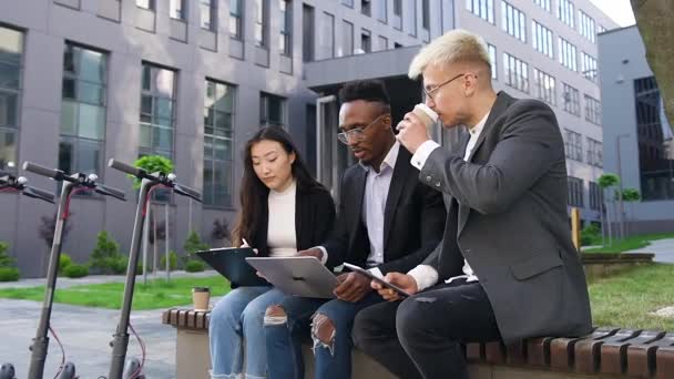 인기있는 긍정적 인 젊은 다종 인종 사무실 직원들 이 커피를 마시는 동안 컴퓨터와 재정 문서를 가지고 도시 건물 근처 야외에서 일하고 있다 — 비디오